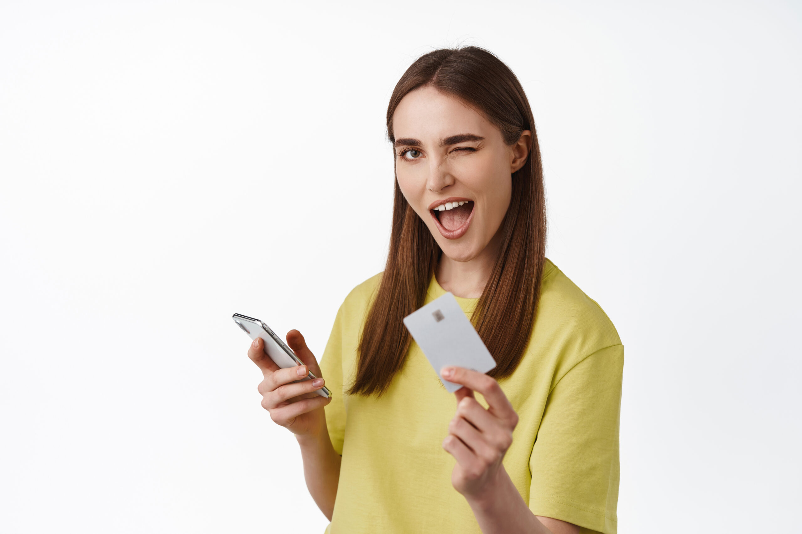 Cheerful girl pays on phone, shopping online with smartphone, showing credit card and winking, got cashback for purchase, standing against white background. Copy space