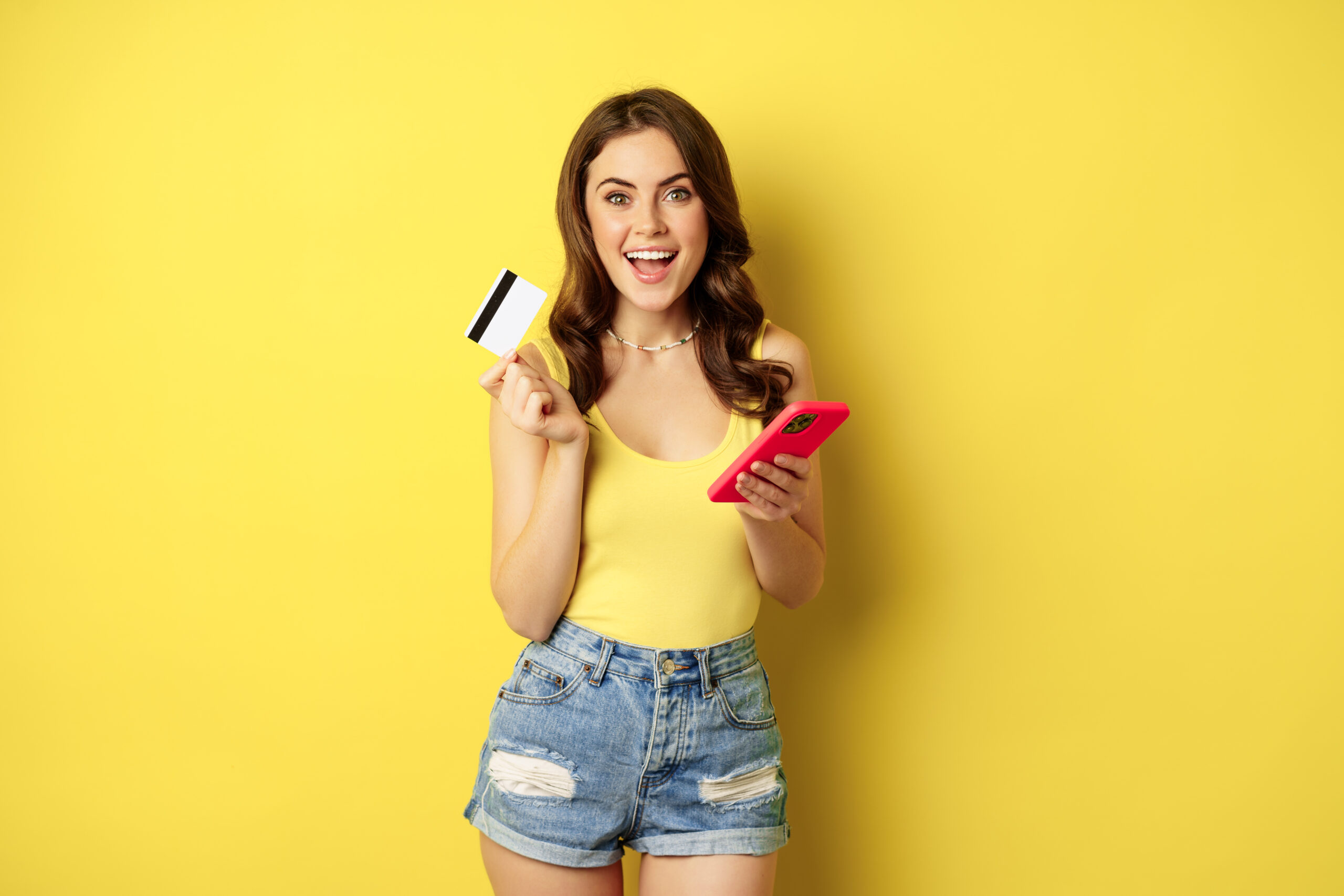 Online shopping. Stylish brunette woman holding smartphone and credit card, paying in app, using mobile phone application, buying smth, standing over yellow background.