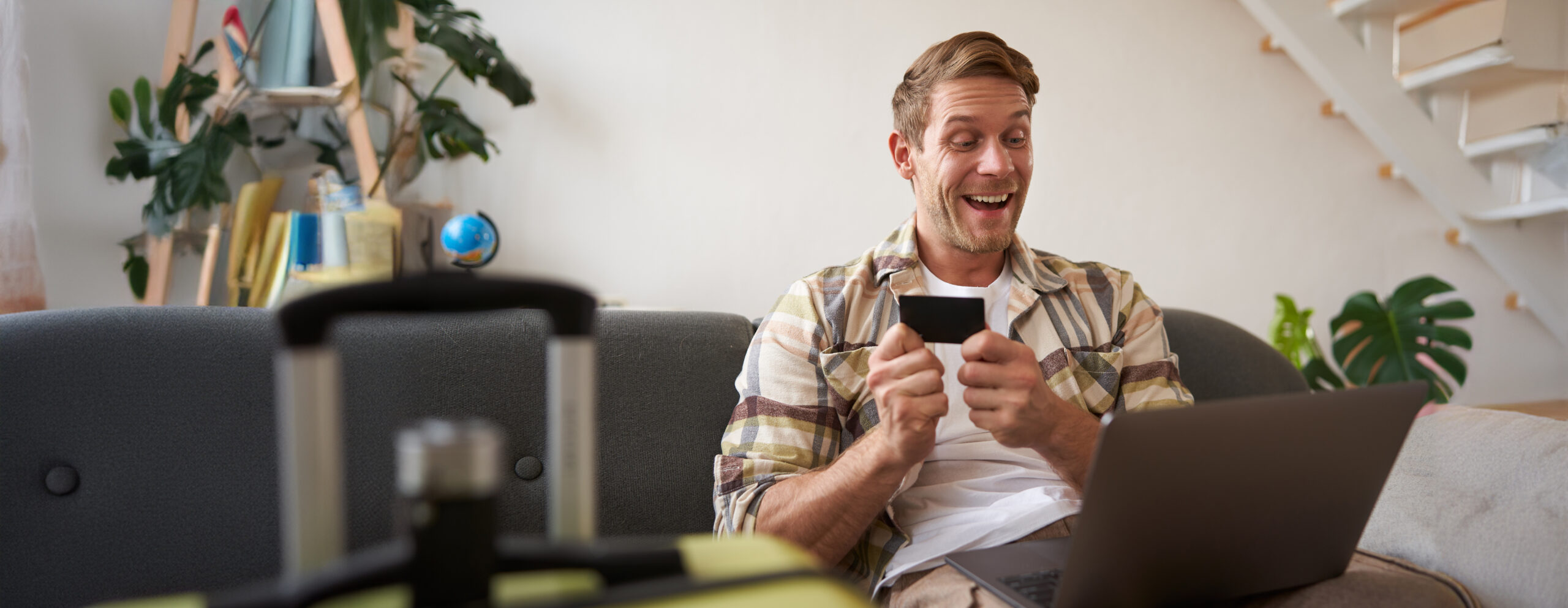 Portrait of happy man, traveller, holding credit card and using laptop, booking vacation, buying tickets online for summer holiday, has suitcase packed.